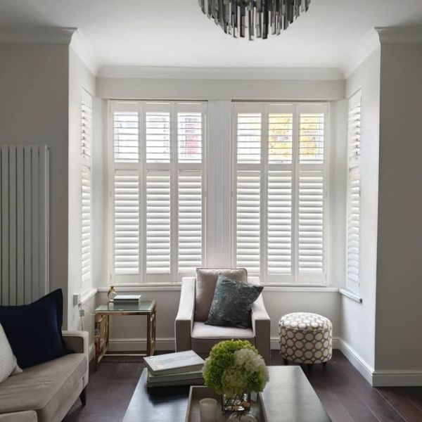 White plantation shutters cover a three-section bay window—two-tier panels with horizontal louvers—partly closed, upper slats slightly open, filtering soft daylight into a neutral living room with an armchair and ottoman.