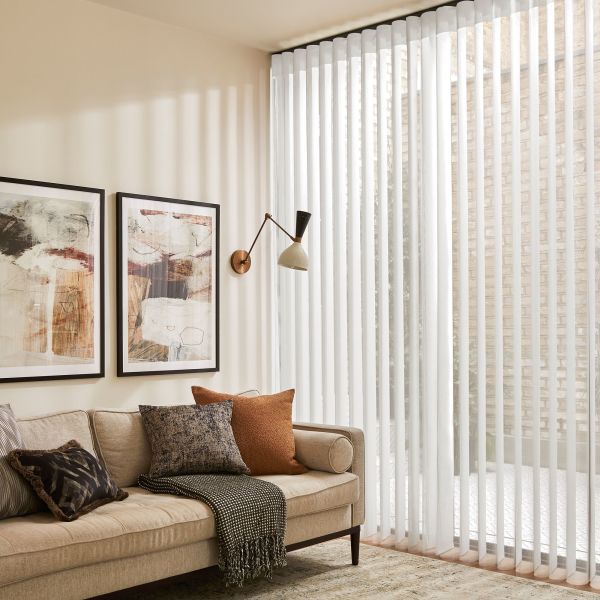 Sheer vertical blinds drawn across a floor-to-ceiling corner window, softly filtering daylight; beige living room with mid-century swing wall lamp, abstract framed art, neutral sofa with textured cushions and throw.