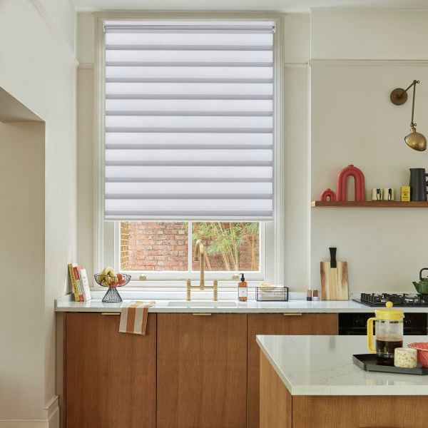 A dual-layer horizontal striped roller blind, lowered three-quarters, filtering daylight. Context: a kitchen with marble countertops, wooden cabinets, brass faucet, and a small brick-wall garden view.