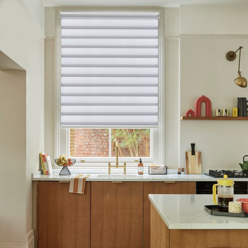 A dual-layer horizontal striped roller blind, lowered three-quarters, filtering daylight. Context: a kitchen with marble countertops, wooden cabinets, brass faucet, and a small brick-wall garden view.