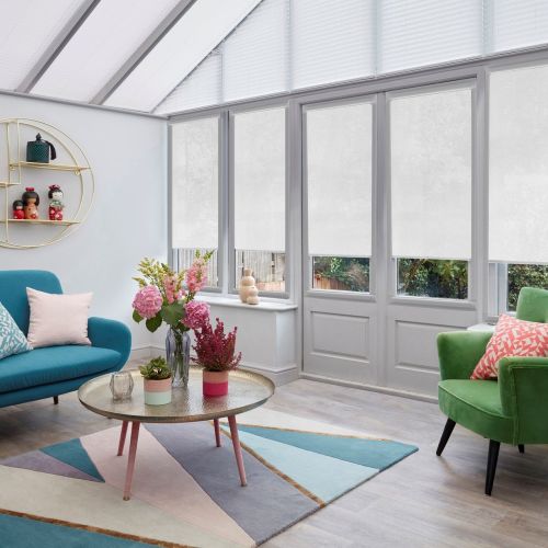 White roller blinds across tall conservatory windows and triangular roof panes, lowered to mid-height and diffusing light; bright sunroom with teal sofa, green armchair, round coffee table, pastel rug.
