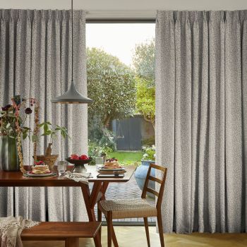Pleated grey patterned curtains, parted at center, framing and admitting soft daylight; wooden dining table set for breakfast beneath a pendant lamp, chairs facing a sunlit garden.