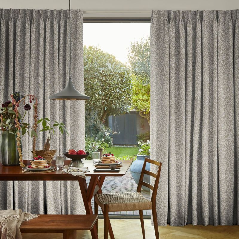 Pleated grey patterned curtains, parted at center, framing and admitting soft daylight; wooden dining table set for breakfast beneath a pendant lamp, chairs facing a sunlit garden.