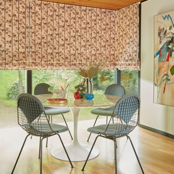 Bamboo-patterned fabric Roman blinds, lowered with soft folds and slightly raised bottom, filtering daylight. In sunlit dining nook with round tulip table, four wire chairs, vases, and garden view.