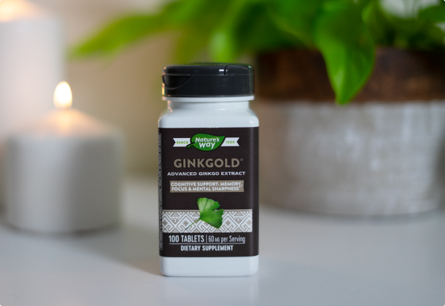 A bottle of Ginkgold sitting on a white table with a lit candle and a leafy plant in the background.