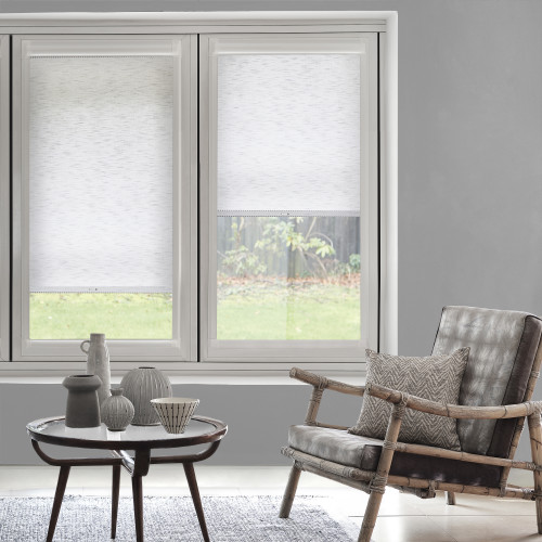 White textured roller blinds, left fully lowered and right partially lowered, filtering daylight over two tall windows; contemporary gray living room with bamboo-frame leather armchair, coffee table, and garden view.