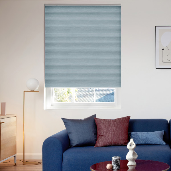 Light-blue cellular (honeycomb) blind, fully lowered with subtle horizontal ribbing, covering most of the window and filtering daylight; small bottom gap reveals rooftops and trees in a modern living room.