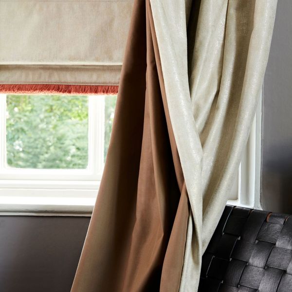 Roman blind with red fringe lowered, flanked by brown blackout and shimmery cream drapes pulled to the side; filtering soft daylight through window onto grey wall and woven leather chair.