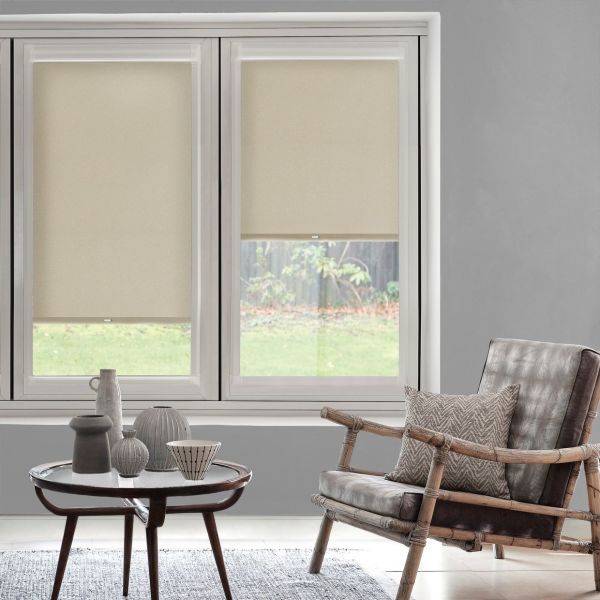 Beige roller blinds, both lowered (left fully, right partially), filtering daylight through two tall windows; muted living room with round wooden table and ceramics, rattan-leather armchair and grassy yard beyond.