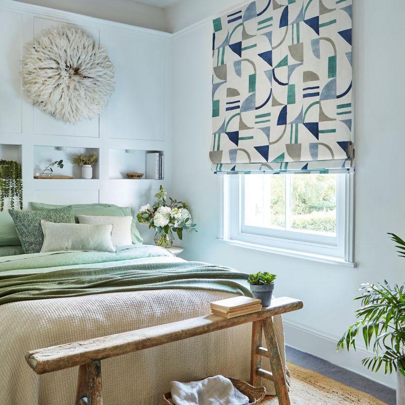 Patterned Roman blind over window, lowered and filtering daylight; geometric teal, blue and gray shapes on pale background. Bedroom setting with pale green bedding, white paneled wall, wooden bench, plants.
