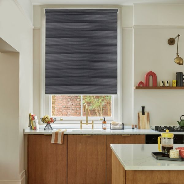 Dark pleated roller blind, fully lowered across a tall window, blocking most light, above a kitchen sink with brass faucet, white countertop, wooden cabinets and a brick courtyard visible below.