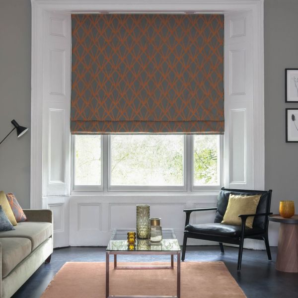 Patterned fabric Roman blind (grey with orange lattice) lowered over bay window, partially filtering bright daylight; living room with grey walls, sofa, black armchair, coffee table and cushions.