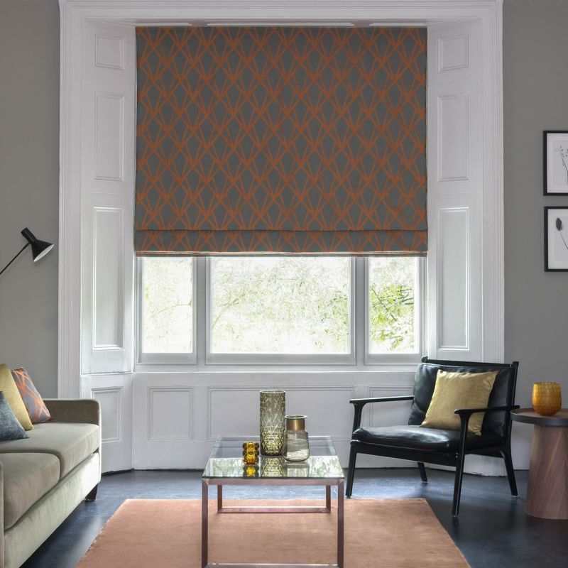 Patterned fabric Roman blind (grey with orange lattice) lowered over bay window, partially filtering bright daylight; living room with grey walls, sofa, black armchair, coffee table and cushions.