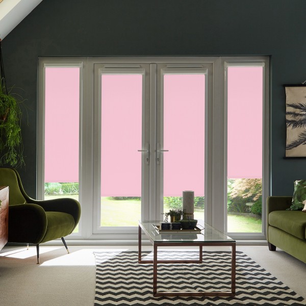 French doors with pink roller blinds lowered halfway, filtering light into a living room with green velvet armchairs, glass coffee table on black-and-white chevron rug, plants against dark teal walls.