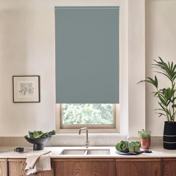 A teal roller blind is partially lowered over a kitchen window, blocking light, above a sink and countertop adorned with a bowl of vegetables, potted plants, and a framed sketch.