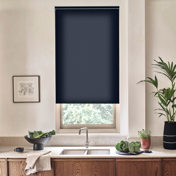 Navy roller blind covering upper portion of a kitchen window, partially lowered; stainless faucet and double sink below on a marble countertop with wooden cabinets, bowl of greens, potted plants.