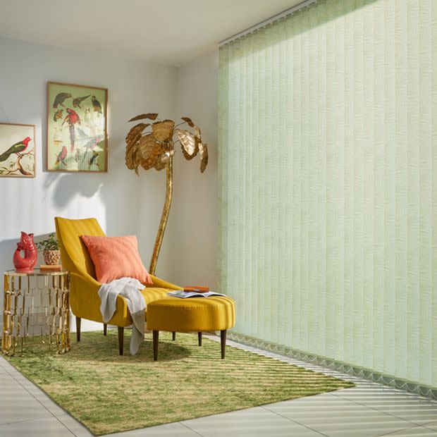 Pale green vertical blinds covering a full-height window, textured and drawn closed, softly diffusing sunlight. Cozy living-room corner with a mustard chaise lounge, coral cushion, gold palm lamp and bird prints.