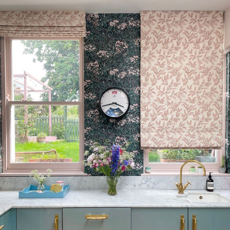 Pair of patterned Roman blindsâ€”left raised, right partially loweredâ€”soft pink botanical fabric filtering daylight over a kitchen sink with marble counter, floral wallpaper and visible green garden outside.