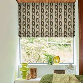 Roman blind with brown‑beige geometric chain pattern, lowered to mid-window, partially filtering daylight; bedroom sill holds green glass vases, potted plant and small fan, with a leafy garden beyond.