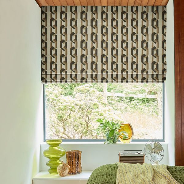 Roman blind with brown‑beige geometric chain pattern, lowered to mid-window, partially filtering daylight; bedroom sill holds green glass vases, potted plant and small fan, with a leafy garden beyond.