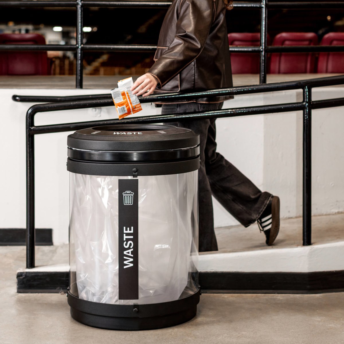 black plastic with a clear body waste bin indoors at a hockey arena with a person walking by