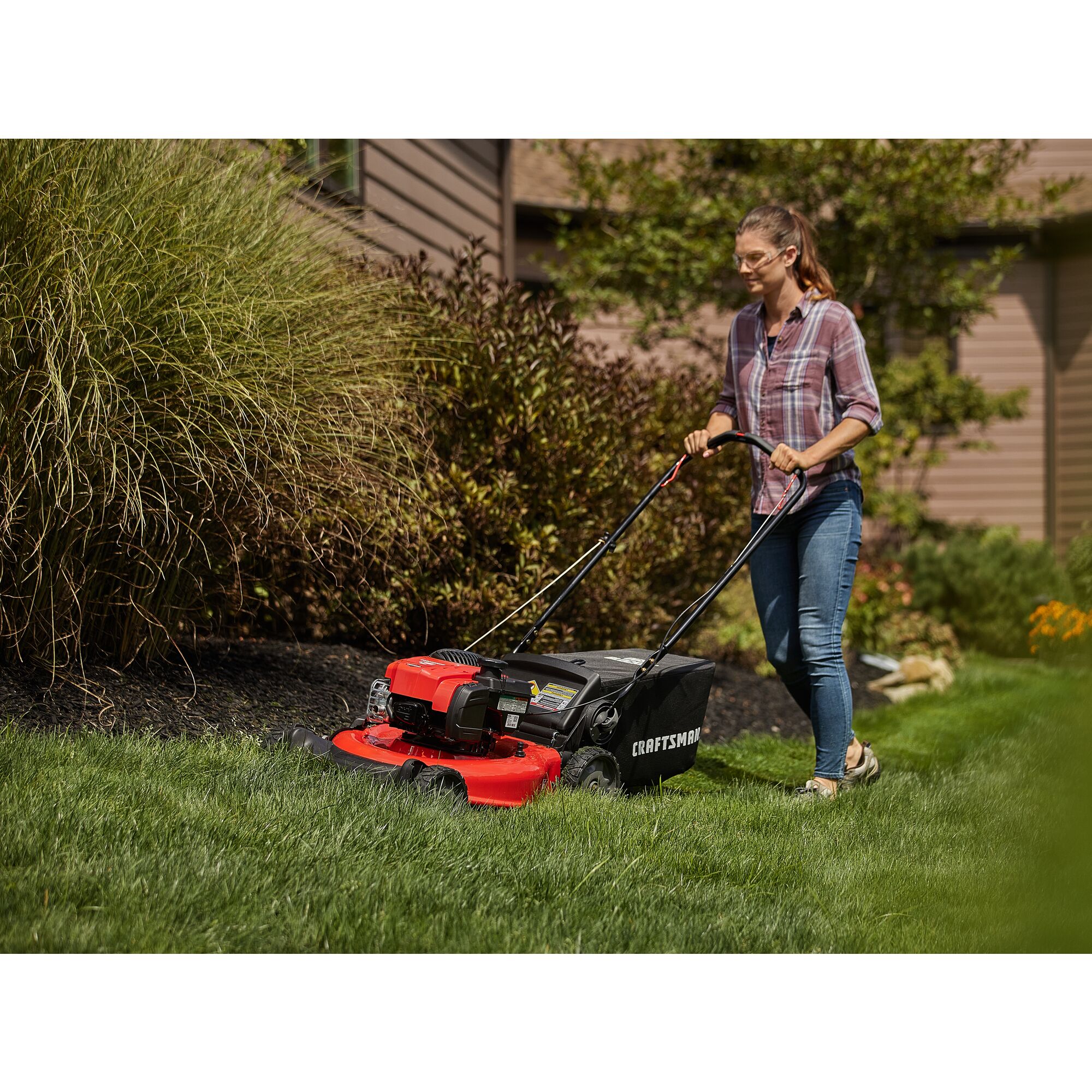CRAFTSMAN 21-in 150cc Push Mower mowing grass around bushes in side view in plaid and jeans