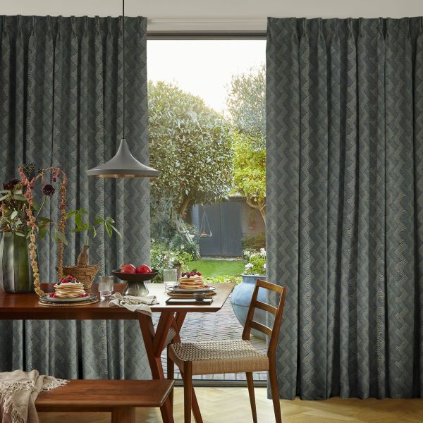 Floor-to-ceiling, patterned pleated curtains, dark blue chevron, partially drawn to frame a sliding glass door; filtering morning light over a dining table set with pancakes, pendant lamp, and backyard garden.