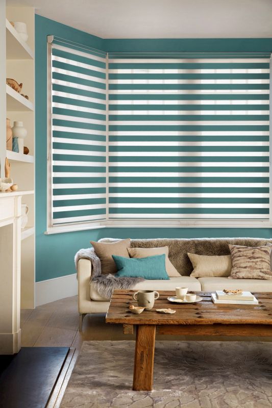 Dual-layer horizontal zebra blinds on a bay window, teal-and-sheer bands, lowered to filter light; set against teal walls above a beige sofa with cushions and a rustic wooden coffee table.