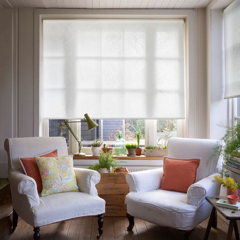 White translucent roller blind with subtle leaf pattern, lowered across a multiâ€‘pane window and gently diffusing daylight; framed by a bright, cozy sitting room with two armchairs, plants, and wooden floor.