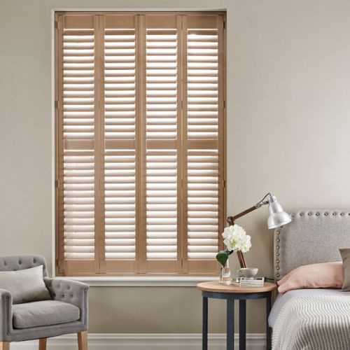 Full-height wooden louvered shutters covering a recessed window, narrow panels with slats tilted to admit diffused light; in a softly lit bedroom beside an upholstered bed, table and chair.