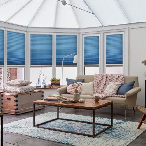 Blue pleated cellular blinds lowered over tall conservatory windows, filtering daylight; beige sofa with knitted throw, patterned cushions and a wooden coffee table on a blue-accent rug form a cozy sunroom.