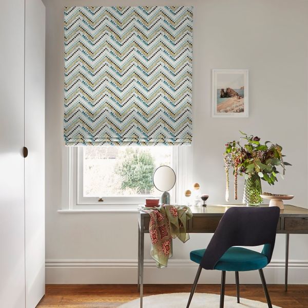Chevron-patterned Roman blind, lowered to mid-window, mosaic teal, gold and black chevrons; filtering soft daylight over a wooden desk with teal chair, vase of flowers and a framed seaside print.