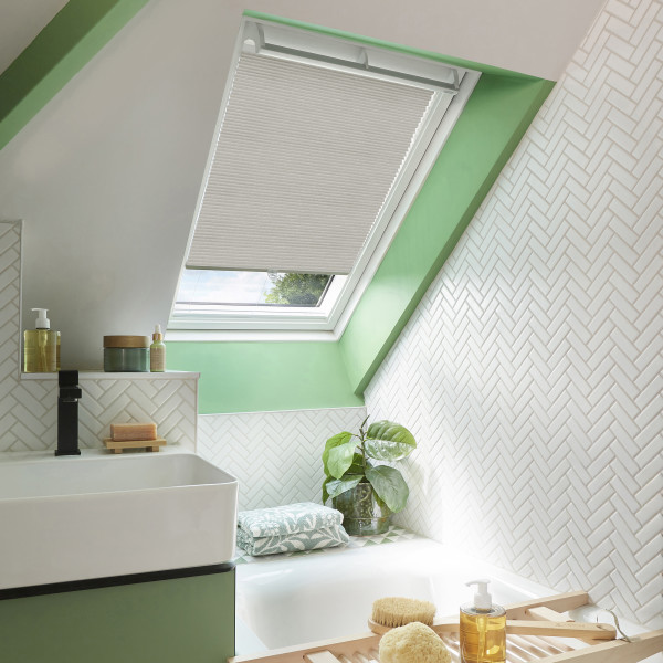 Beige pleated skylight blind, lowered over most of the roof window leaving a small bottom gap, filtering light into a bright bathroom with mint trim, white herringbone tiles and bathtub.