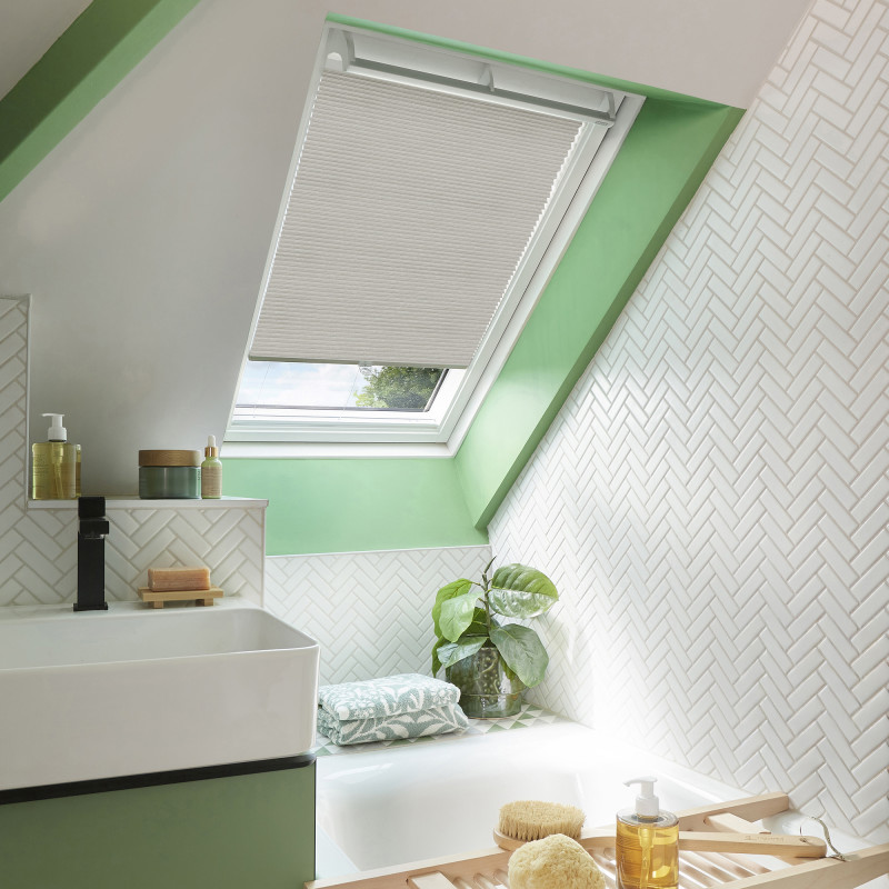 Beige pleated skylight blind, lowered over most of the roof window leaving a small bottom gap, filtering light into a bright bathroom with mint trim, white herringbone tiles and bathtub.