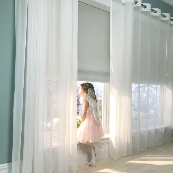 Sheer grommet-top curtains over a lowered roller blind, panels parted, filtering soft daylight as a girl in a dress and wings leans at the sill, bright room with garden view.