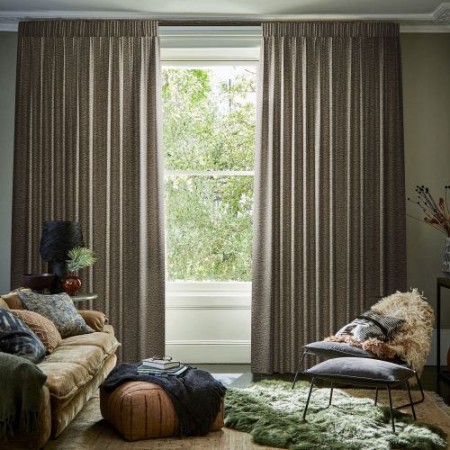 Heavy pleated brown curtains, hung full-length on a concealed track, parted to frame the window; filtering daylight. Living room setting with sofa, ottoman, plush rug and greenery visible outside.