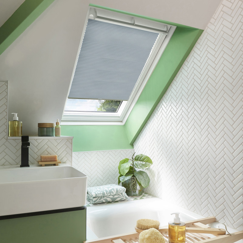 A light-gray pleated skylight blind, partially lowered and filtering daylight, in a sloped bathroom with a green-framed skylight, white herringbone tiles, bathtub, sink, plant, and toiletries.