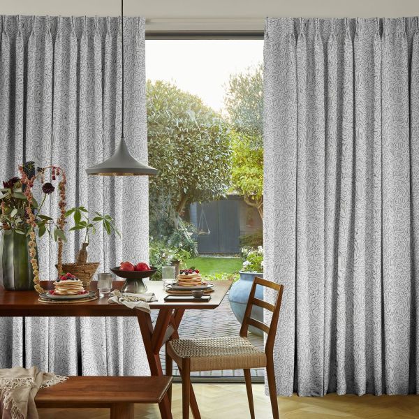 White pleated curtains with abstract squiggle print, parted to frame the door and filtering soft daylight into a cozy dining area with wooden table, pendant lamp, and leafy garden beyond.