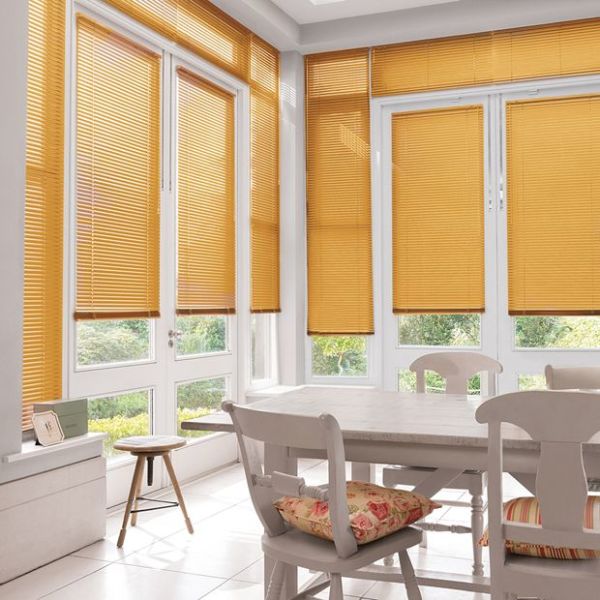 Honey‑orange horizontal slatted blinds cover tall bay windows and French doors, mostly lowered and filtering sunlight; bright dining area with white table, chairs with floral cushions and garden view.