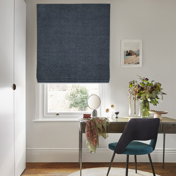 Dark-blue textured Roman blind lowered above the sill, filtering daylight over a window; small home desk scene with teal chair, mirror, vase of flowers and framed seaside print.