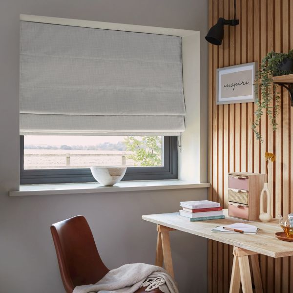 Light grey Roman blind, lowered and softly folded, filtering daylight through a grey-framed window; home office corner with wooden slat wall, desk, books, bowl on sill, leather chair and trailing plant. Framed print reads: "inspire."