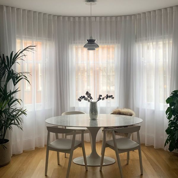 Sheer white floor-to-ceiling curtains on a curved bay of three windows, gently diffusing daylight; round marble-topped dining table with chairs and vase on a warm wooden floor.