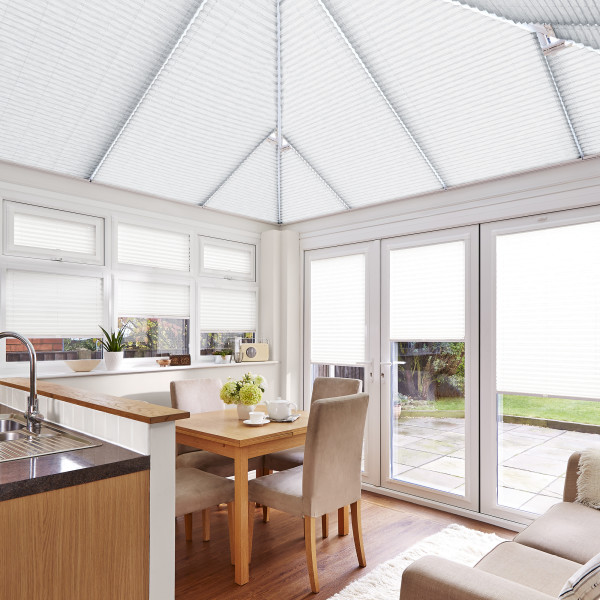 Object: white pleated blinds covering windows, French doors and pitched conservatory roof. Action: lowered, filtering and diffusing bright daylight. Context: airy kitchen-dining conservatory with wooden table and patio garden view.