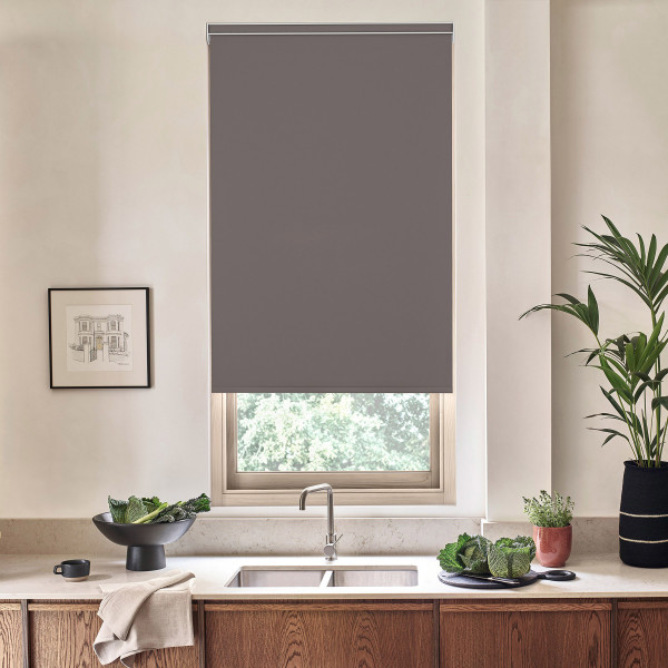 Gray roller blind hangs lowered over a window, revealing treetops below; stainless faucet and double sink sit on a marble countertop with bowls of leafy greens and potted plants.