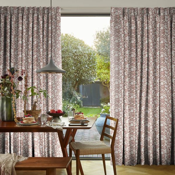 Full-length pleated curtains with a pink squiggle pattern, parted to reveal sliding glass door and gently filtering daylight; framing a wood dining table set for breakfast and sunlit garden beyond.