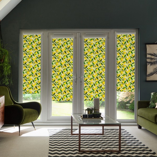 Four tall window panels fitted with yellow-and-green leaf-patterned roller blinds, filtering sunlight into a living room with green armchairs, glass coffee table, and a chevron rug.