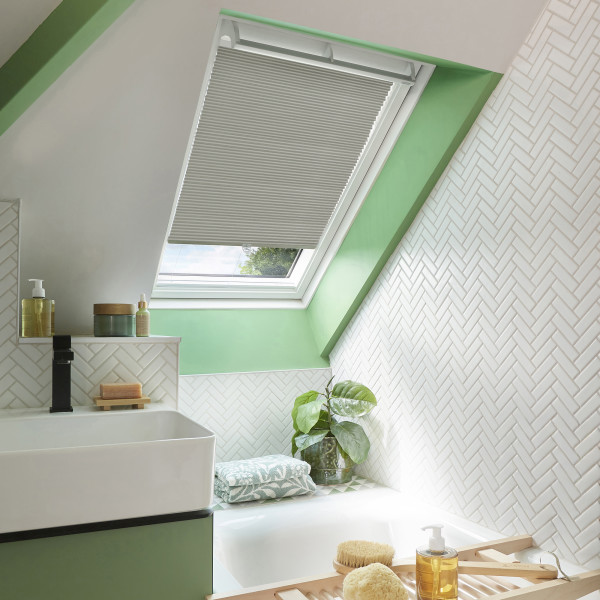 A light-gray pleated skylight blind, partially lowered across a slanted roof window, filtering daylight; attic bathroom with mint-green trim, white herringbone tiles, bathtub, sink, potted plant and toiletries.