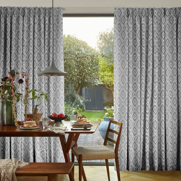Gray full-length pleated curtains with white diamond pattern, hung on a sliding glass door, drawn partly open to reveal a sunlit garden; dining area with wooden table, chair, pendant lamp.