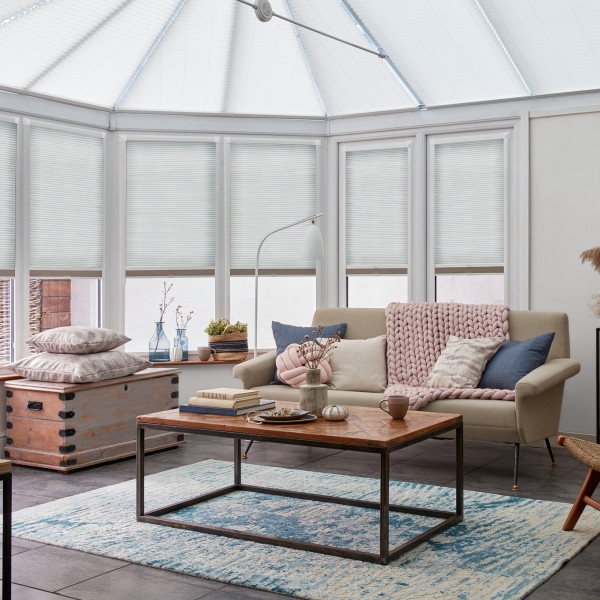 White pleated blinds on bay windows and conservatory roof, drawn to diffuse light; filtering soft daylight into a sunroom-style living area with neutral sofa, pink chunky throw, and wooden coffee table.