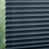 Dark gray pleated cellular blinds, stacked closed with visible cords and staples, blocking light; filtering soft daylight from a blurred green outdoor view through a window, creating diffuse interior lighting.